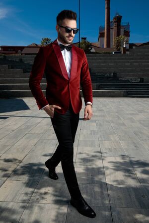 side view of confident young man wearing red velvet tuxedo and sunglasses, walking and holding hand in pocket, outdoor in an urban scene; full bodyの写真素材