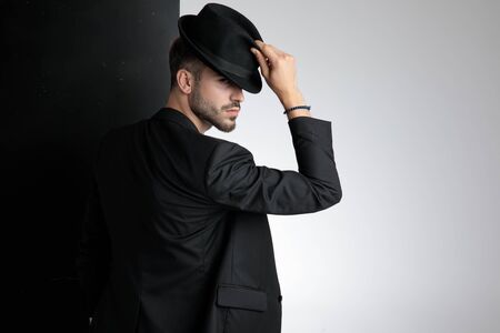 back view of polite young man looking to side and holding black hat, standing on black and white background in studioの写真素材