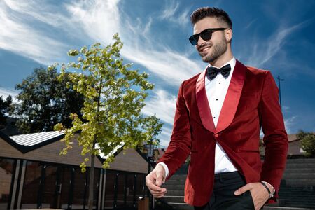 side view of optimistic young man wearing red velvet tuxedo and sunglasses, smiling and walking, holding hand in pocket, outdoor in an urban sceneの写真素材