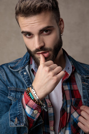 seductive young man wearing blue denim jacket, touching lips and standing on brown background in...