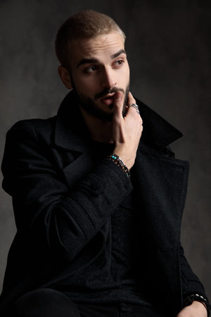 attractive fashion model wearing longcoat, holding hand to cheek, sitting on grey background in...