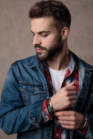 casual young fashion man wearing blue denim jacket, adjusting bracelets and looking down side, on...