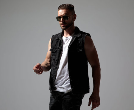 Determined man walking while wearing sunglasses and a black jeans vest on gray studio background