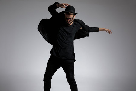 handsome casual man wearing a hat and glasses dancing around with passion against gray studio...