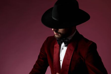 close up of young fashion model wearing red velvet tuxedo and black hat, looking to side and hiding behind hat on pink background in studioの写真素材