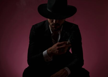 Determined mysterious man smoking and leaning on his leg while wearing a black suit and hat, sitting on cherry-colored studio backgroundの写真素材