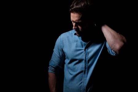 mysterious casual man wearing blue shirt standing in the shadows and scratching his head pensive against black studio backgroundの写真素材