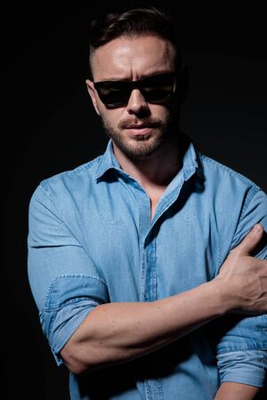 good looking casual man wearing blue shirt and sunglasses standing with hand on arm and staring at camera serious against black studio backgroundの写真素材