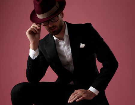 Mysterious man taking off his glasses and leaning on his leg while wearing tuxedo, a red hat, sitting on pink studio backgroundの写真素材