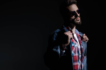 Motivated young man adjusting his jacket and looking away while wearing sunglasses and a checkered shirt, standing on black studio backgroundの写真素材