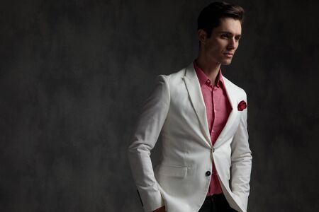 Hopeful elegant man looking away with both hands in his pockets while wearing a white suit and pink shirt, standing, on gray wallpaper studio backgroundの写真素材