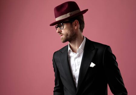 Confident elegant man looking away while wearing tuxedo, a red hat and glasses, standing on pink studio backgroundの写真素材