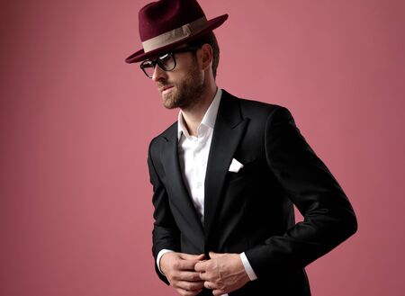 Hopeful elegant man looking away and unbuttoning his jacket while wearing tuxedo, a red hat and glasses, standing on pink studio backgroundの写真素材