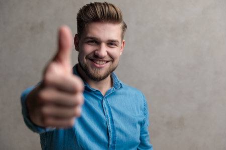 Positive casual man smiling and giving a thumbs up while wearing a blue shirt, standing on gray...