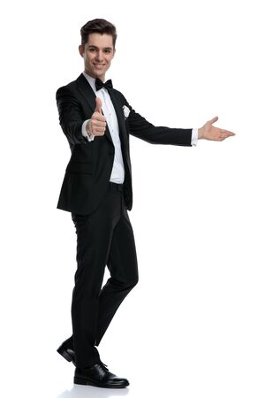 side view of confident young man in tuxedo making thumbs up sign and inviting to side, walking isolated on white background, full bodyの写真素材