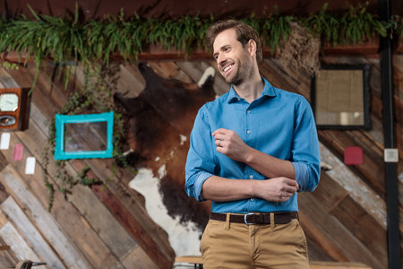 Cheerful casual man laughing and looking away while wearing a blue shirt and brown pants, standing on a coffeeshop backgroundの写真素材