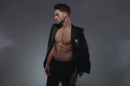 young fashion model holding jacket on shoulders and looking down, standing on grey backgroundの写真素材