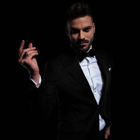 attractive businessman wearing black tuxedo standing with one hand in pocket and gesturing with one hand raised on black studio backgroundの写真素材
