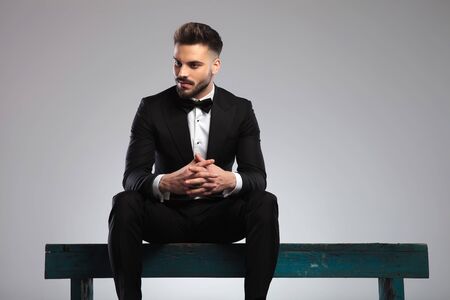 Eager young model looking away and smiling while wearing tuxedo, sitting on a bench on gray studio backgroundの写真素材