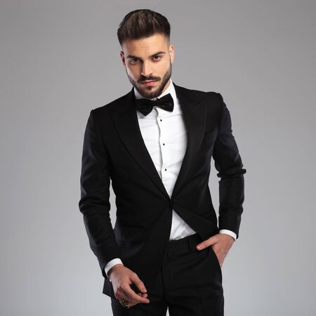 Confident young model holding his hand in his pocket while wearing tuxedo, standing on gray studio backgroundの写真素材
