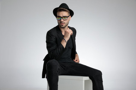 sexy cool guy wearing hat and sunglasses, holding elbow on knee and looking to side, sitting on grey...
