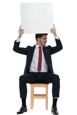 elegant businessman holding empty board above head, looking to side and sitting isolated on white backgroundの写真素材