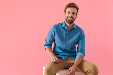 happy casual man in denim shirt smiling and holding elbow on thigh, sitting on pink background