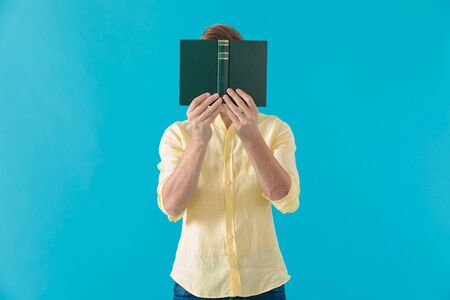 Young casual man hiding his face behind a book, standing on blue studio backgroundの写真素材
