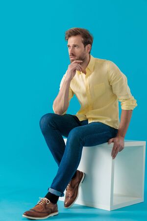 Thoughtful casual man looking away with his hand on his chin, wondering and solving his problems, sitting on blue studio backgroundの写真素材