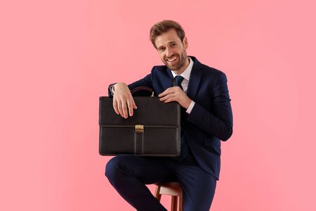 Positive businessman holding a briefcase and laughing while sitting on a stool on pink studio backgroundの写真素材