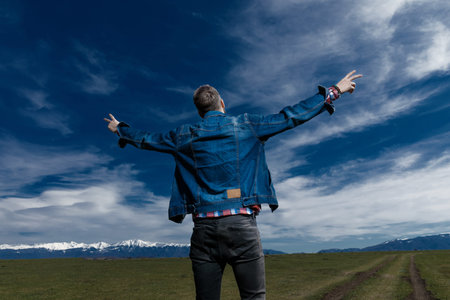 Rear view of a young man gesturing peace with both hands while wearing jeans jacket and standing on...
