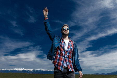 Handsome young man holding his hand in the air while wearing jeans jacket and sunglasses, standing...
