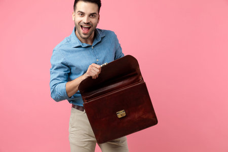 excited young man laughing and opening suitcase on pink background
