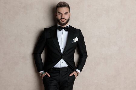 Bothered groom looking forward with both hands in his pockets while wearing tuxedo and standing on wallpaper studio backgroundの写真素材