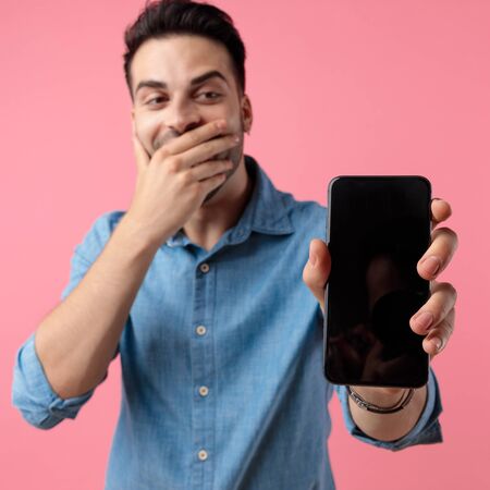 shocked young casual man covering his mouth while presenting the screen of his mobile phone on pink backgroundの写真素材