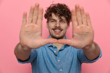 young casual guy in denim shirt smiling and holding hands, framing, standing on pink background