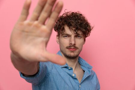 sad casual guy in denim shirt holding hand in and stopping, standing on pink backgroundの写真素材