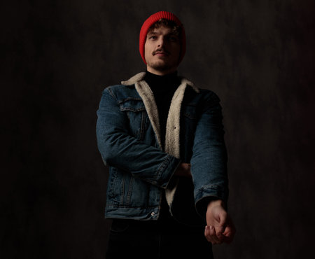 dramatic cool model wearing red hat and denim jacket, holding arm in a fashion pose on black...