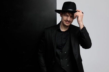 Positive fashion model fixing his hat and greeting while wearing suit and standing on black and...