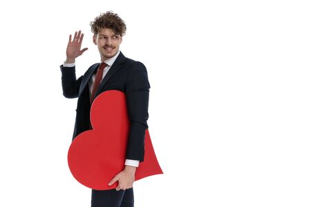 Happy businessman holding heart shape, smiling and waving while wearing suit and standing on white studio backgroundの写真素材