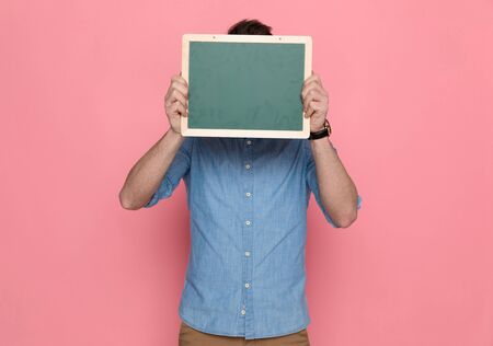casual man hiding his face behing a board, wearing a blue shirt and standing on pink studio backgroundの写真素材
