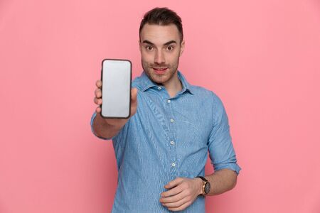 sexy young casual man showing his telephone to the camera, standing and wearing a blue shirt on pink studio backgroundの写真素材