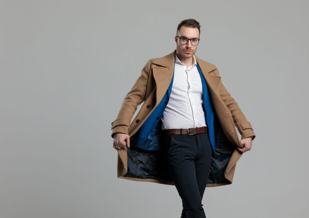 attractive young guy wearing glasses and opening long coat, standing on grey background