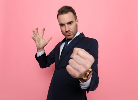 upset young man in suit holding fist up, pointing finger and threatening, standing on pink...
