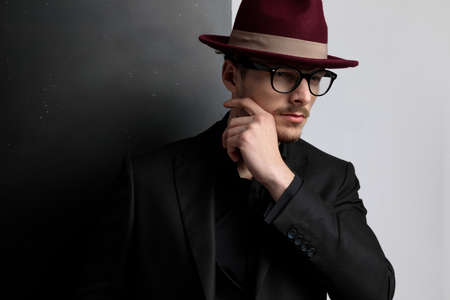 dramatic portrait of an elegant fashion guy touching his face, looking away and wearing a burgundy hat on black and white studio backgroundの写真素材