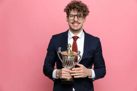 excited young man in navy blue suit wearing glasses, holding trophy and smiling on pink backgroundの写真素材
