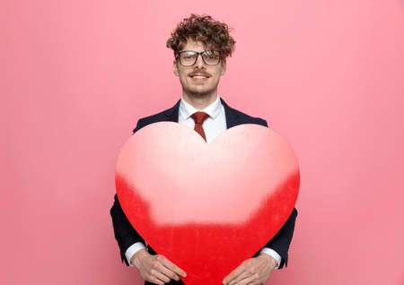 happy young businessman in suit holding big red heart and smiling on pink backgroundの写真素材