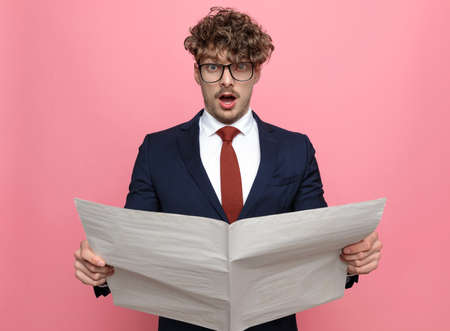 shocked young man in navy blue suit wearing glasses, reading newspaper and opening mouth, posing on pink backgroundの写真素材