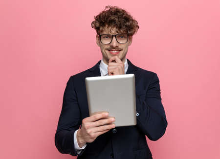 happy young businessman in navy blue suit wearing glasses holding hand to chin and thinking, holding hand and smiling on pink backgroundの写真素材