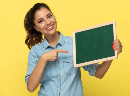 Young casual woman holding and pointing at blackboard, smiling while standing on yellow studio backgroundの写真素材